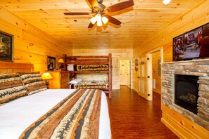 A log cabin-style bedroom with a large bed, a stone fireplace, a ceiling fan, and a television mounted above the fireplace. - Luxury 14br Haven Views Games Hot Tubs More (Sevierville)
