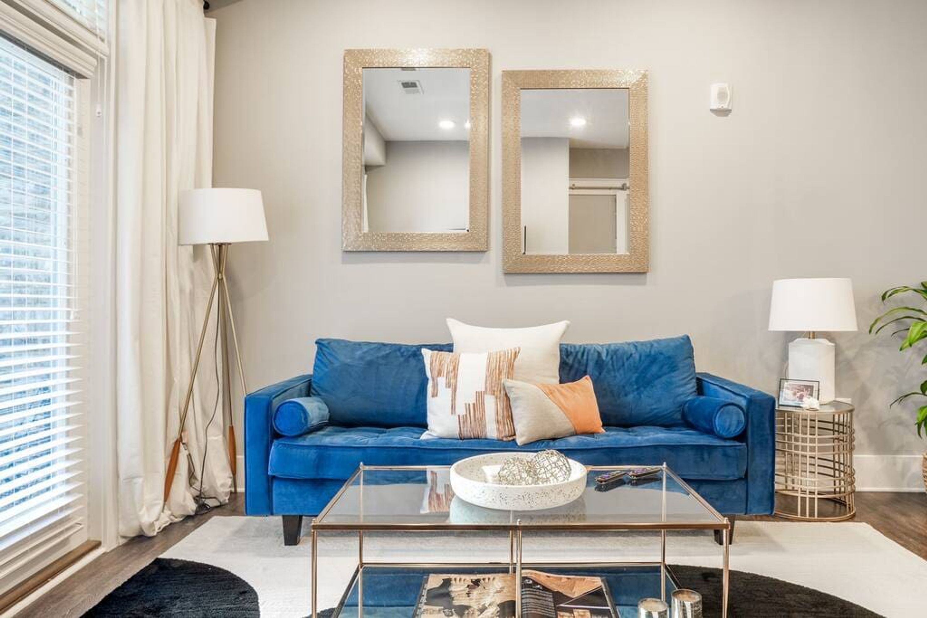 Un salón con un sofá azul, dos espejos en la pared, una mesa de centro de cristal y una mesita de noche con una lámpara.
