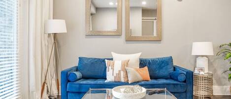Un salón con un sofá azul, dos espejos en la pared, una mesa de centro de cristal y una mesita de noche con una lámpara.