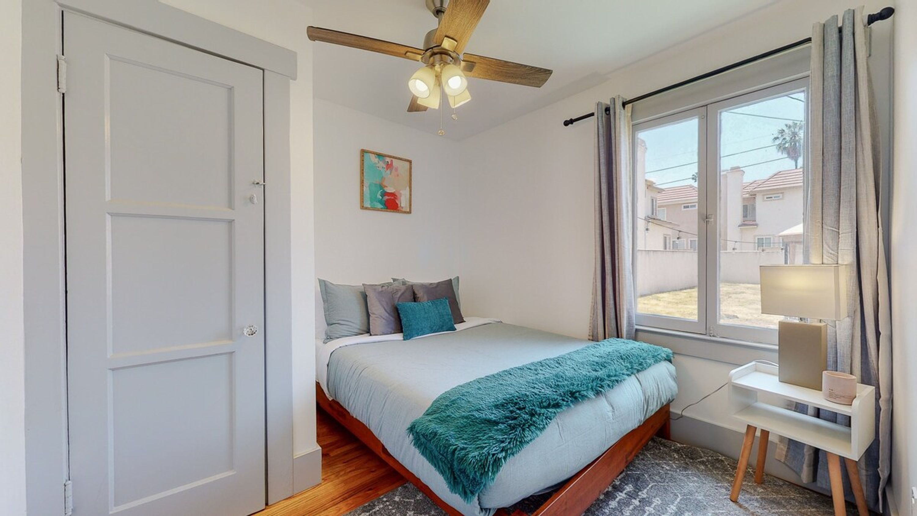 A bedroom with a bed, a ceiling fan, a window with curtains, and a small bedside table with a lamp.