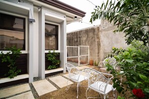 Un patio con sillas de metal blanco y un portón blanco, rodeado de plantas y un muro de hormigón.