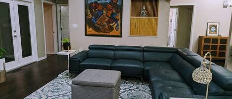 A living room with a dark green sectional sofa, a grey ottoman, and a patterned rug. There is a painting on the wall and a small statue in a wooden display.