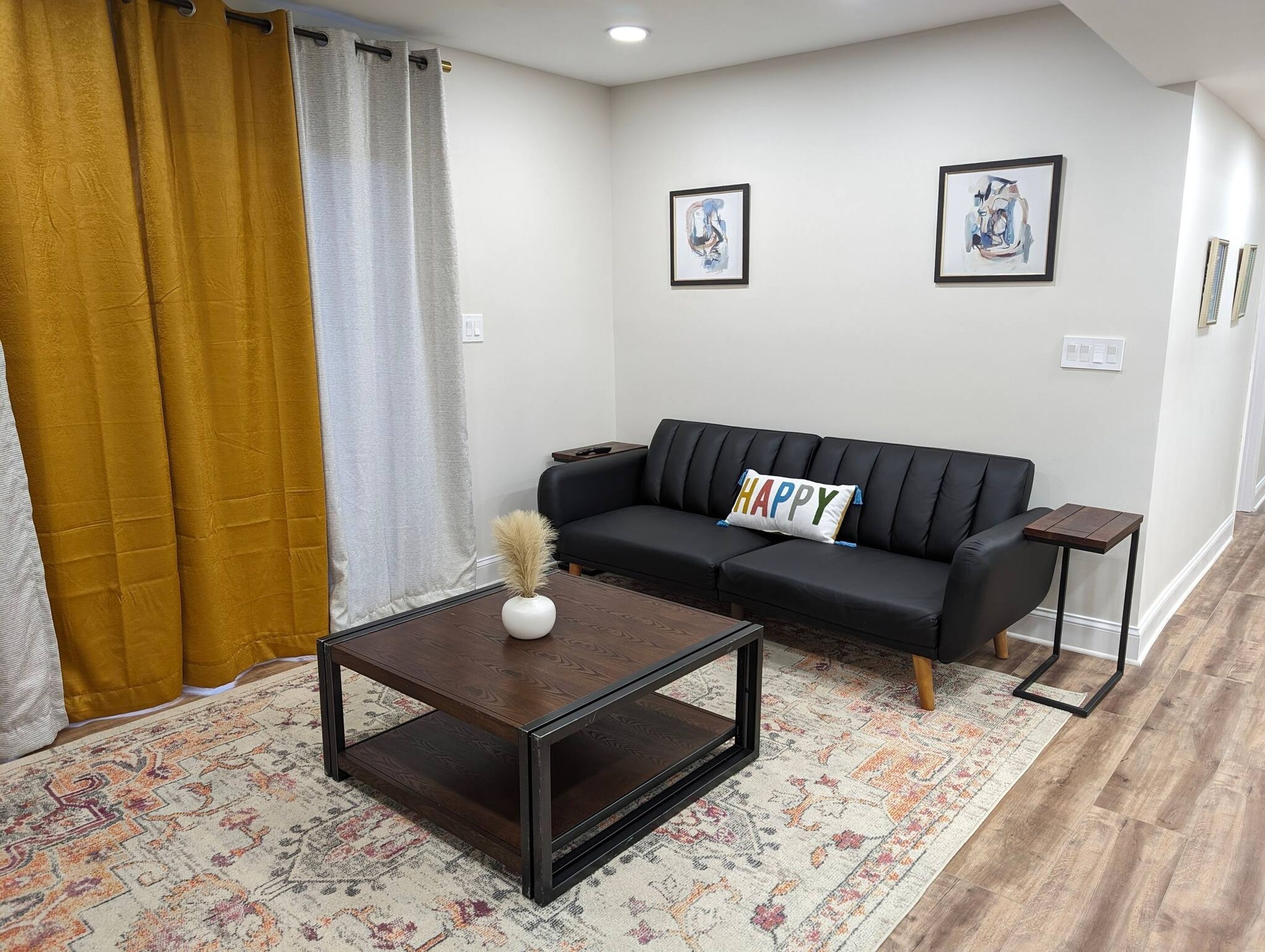 Uma sala de estar com um sofá preto, uma mesa de centro, um tapete estampado e duas obras de arte emolduradas na parede.