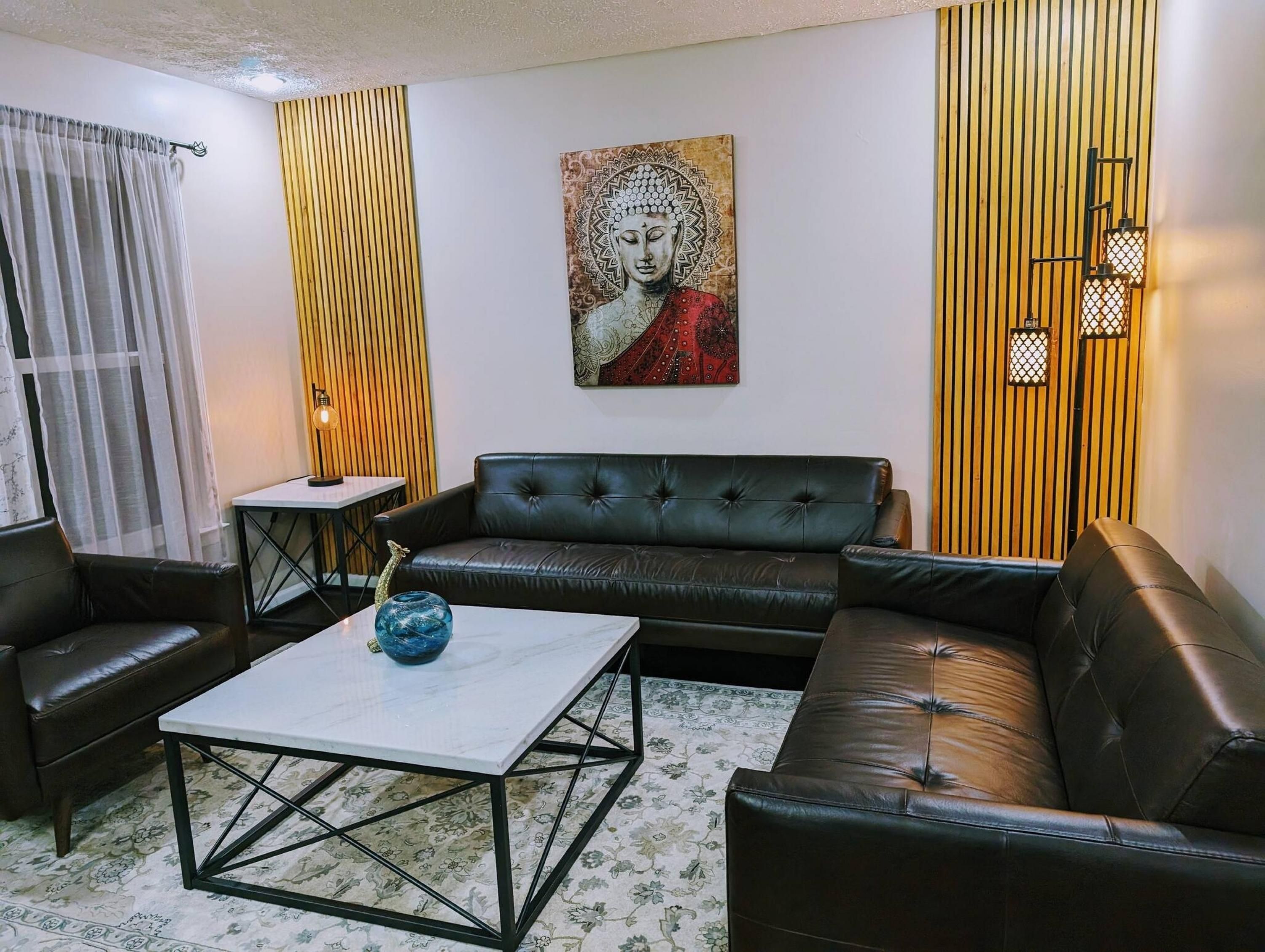 A living room with a black leather sofa, a white coffee table with a blue vase, a small side table, and a wall-mounted lamp.