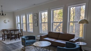 A spacious living room with a brown sofa, two blue armchairs, a round coffee table, and a dining area with a wooden table and chairs. Large windows provide natural light and views of trees. - 2756 Bridgegate Trce NE GA - Basement (Marietta)