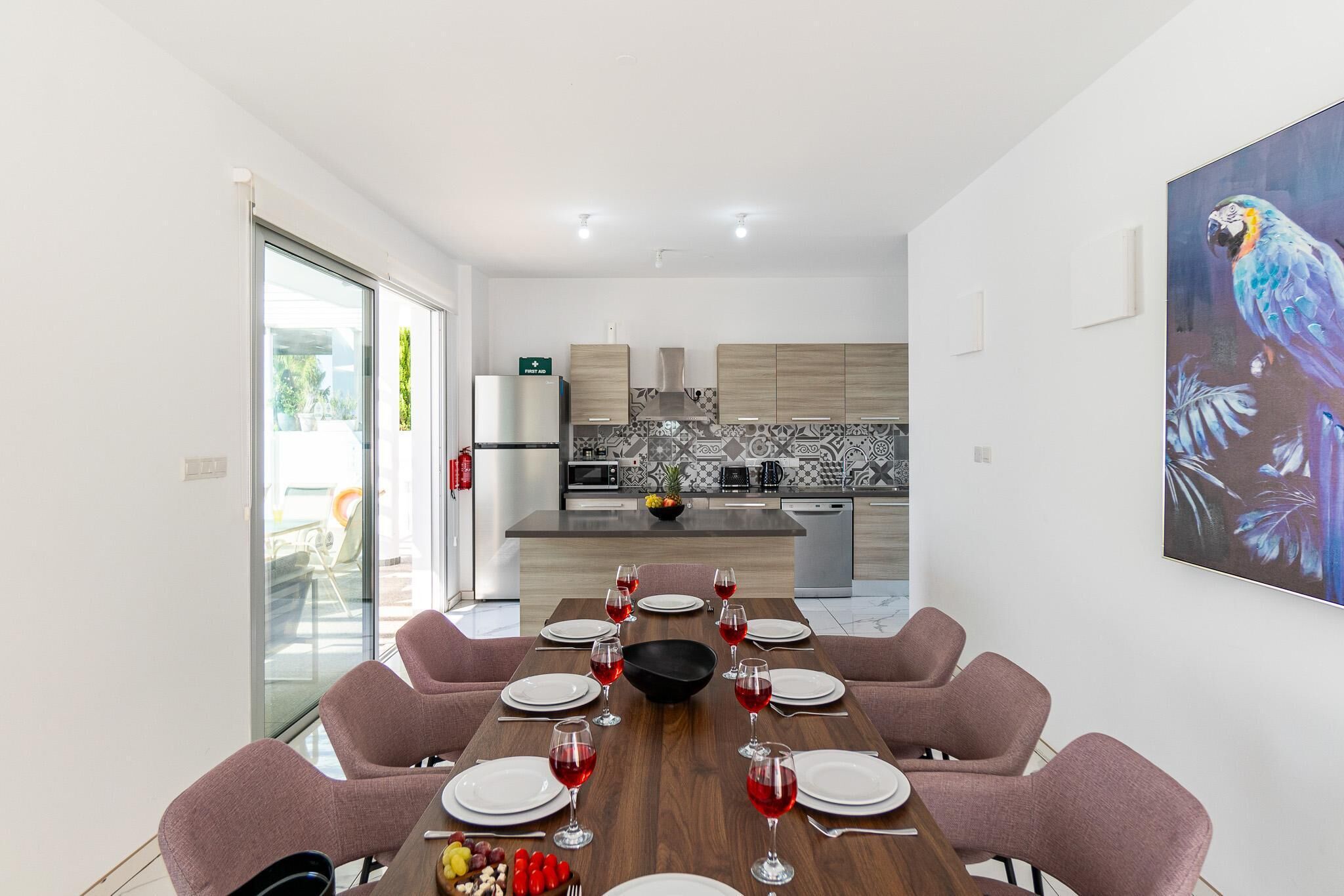 Un moderno comedor con una larga mesa dispuesta para seis, una cocina al fondo y un cuadro grande de un loro en la pared.