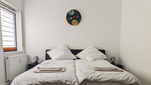 Een tweepersoonsbed met witte lakens, twee nachtkastjes met lampen, een radiator en een aan de muur hangend kunstwerk.
