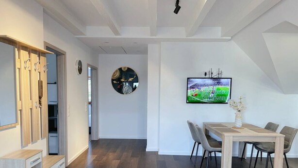Een moderne eetruimte met een houten tafel en stoelen, een tv aan de muur en een hal met een spiegel en houten kasten.