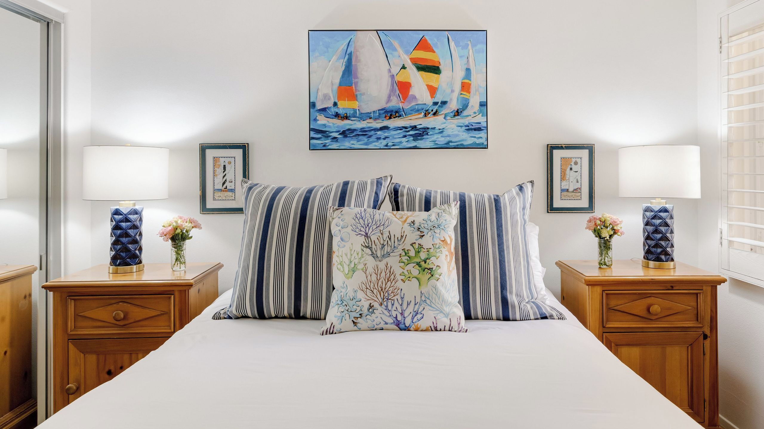 A hotel room with a bed, two nightstands, a painting of sailboats, and a window with blinds.