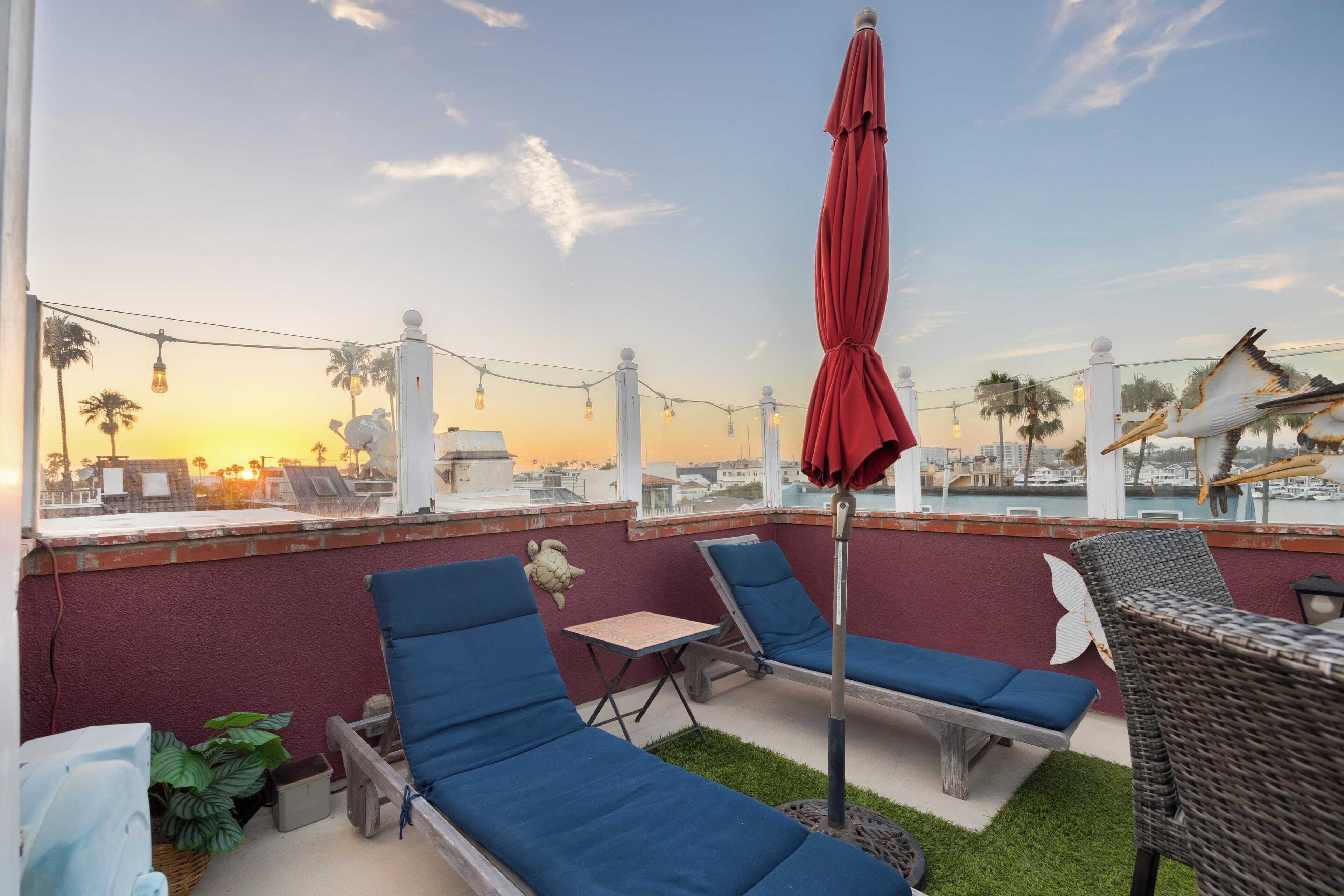 Una terraza en la azotea con sillones azules, una sombrilla roja y vistas al atardecer sobre la ciudad.