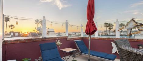 Una terraza en la azotea con sillones azules, una sombrilla roja y vistas al atardecer sobre la ciudad.