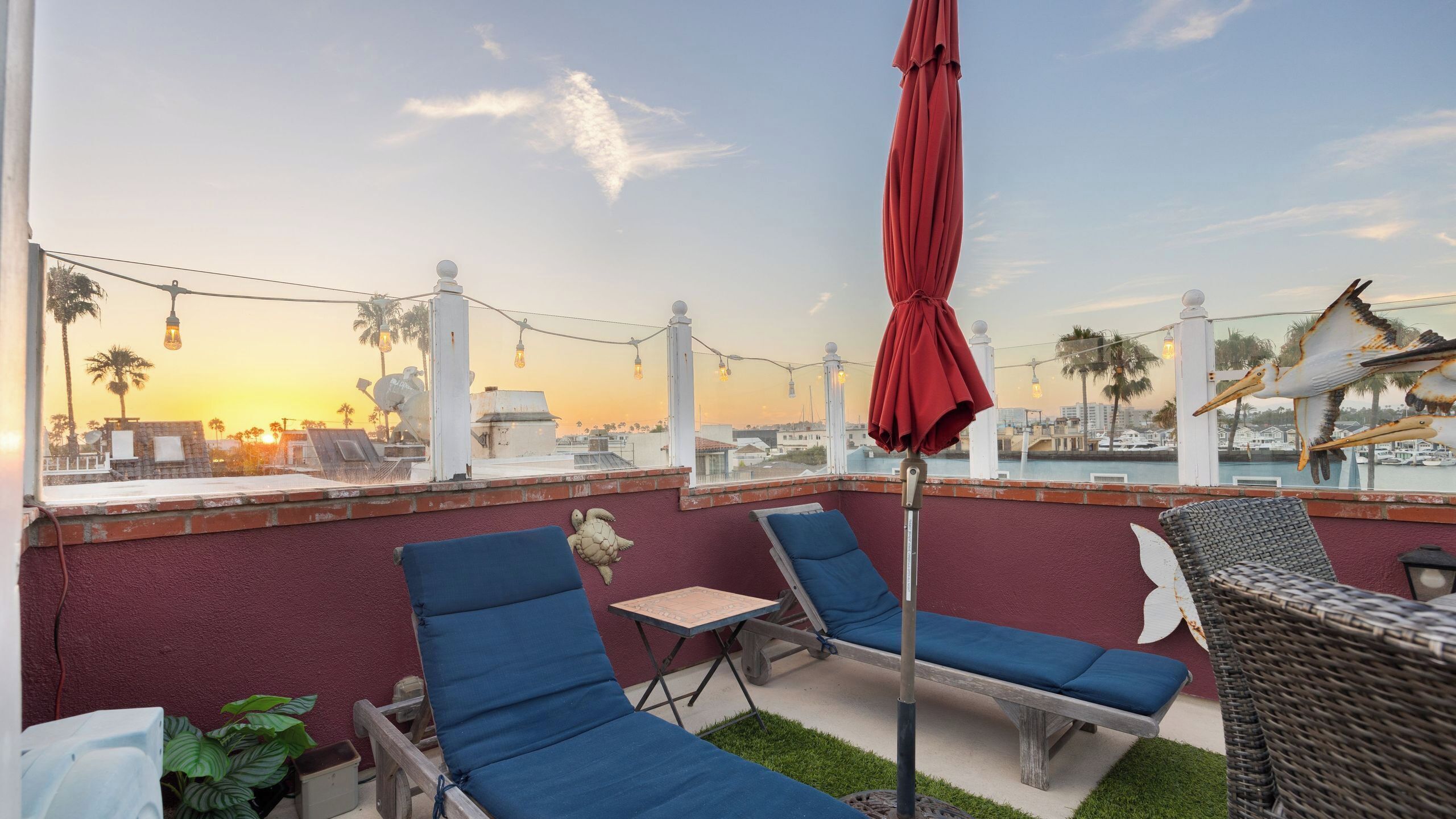 Eine Dachterrasse mit blauen Liegestühlen, einem roten Sonnenschirm und Blick auf den Sonnenuntergang über einem Yachthafen.