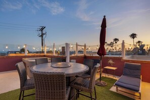 A rooftop terrace with a round table, wicker chairs, a small table, a lounge chair, and string lights. - Shoreline Villa by Avantstay Steps to the Beach, Bay, & Pier! (Newport Beach)