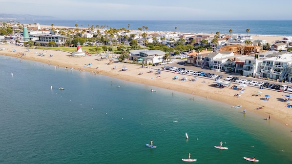 An aerial view of a coastal town with a beach, a lighthouse, and a marina with boats.