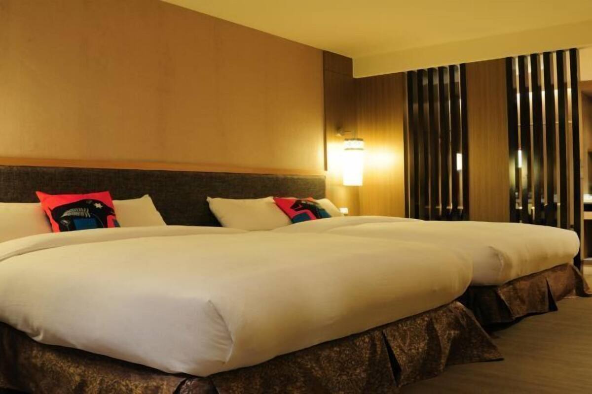 A hotel room with two beds, a headboard, a wall-mounted lamp, and a vertical wooden panel.