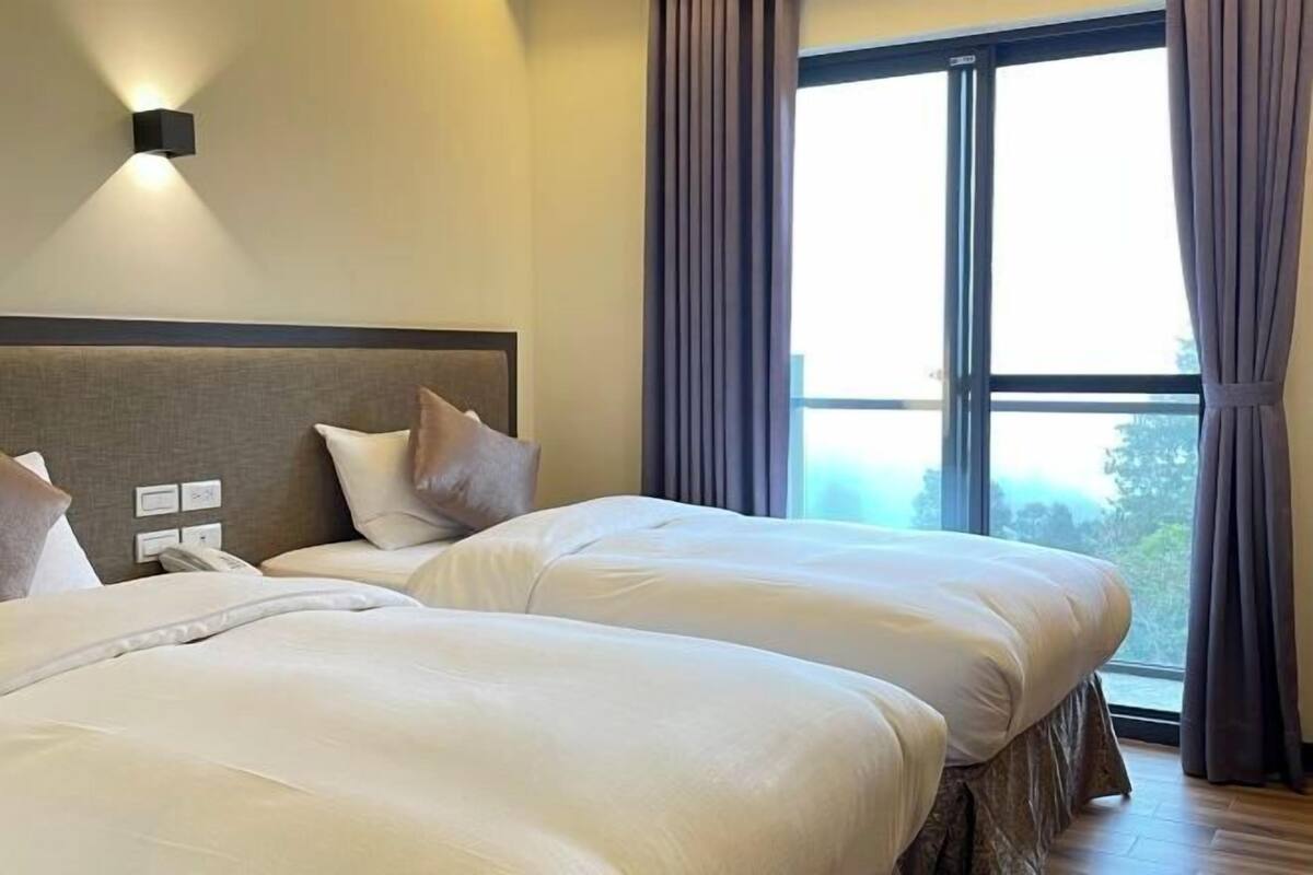 A hotel room with two beds, a headboard, a bedside telephone, and a large window with curtains.