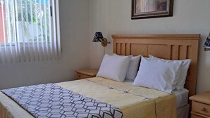 Un dormitorio con cama, dos mesitas de noche, una lámpara de noche, una ventana con persianas y un cuadro enmarcado en la pared.