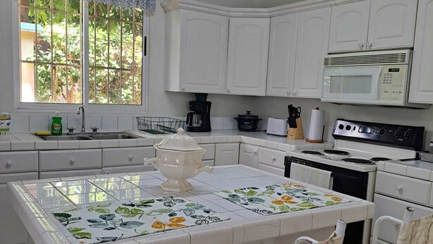 Una cocina con gabinetes blancos, una isla central con mantel floral, un microondas, una estufa y una ventana con vistas a follaje.