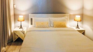 Uma cama bem arrumada com duas luminárias de cabeceira, uma cabeceira e dois travesseiros decorativos.