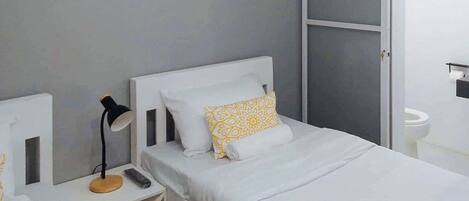 A small, neatly arranged bedroom with a single bed, a bedside table with a lamp, and a mirror.
