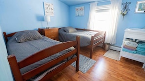A bedroom with two wooden beds, a bedside table, a lamp, a wall art, a window with curtains, and a white dresser with towels. - Sunset Paradise Cottage (Aurora)
