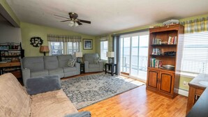 A living room with a sofa, armchairs, a rug, a ceiling fan, a bookshelf, and a view through a glass door. - Sunset Paradise Cottage (Aurora)