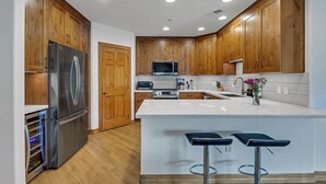 A modern kitchen with wooden cabinets, stainless steel appliances, a central island with bar stools, and a white backsplash. - Riverfront Luxury- Minutes to Skiing+Garage+Walk to shops & restaurants! (Edwards)