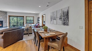 A dining area with a wooden table set for four, surrounded by leather chairs. A brown leather sofa is positioned to the left, and a fireplace is visible in the background. - Riverfront Luxury- Minutes to Skiing+Garage+Walk to shops & restaurants! (Edwards)