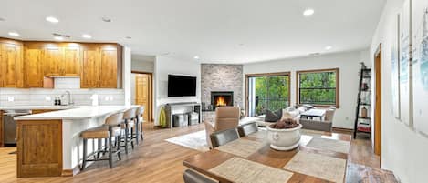 A modern kitchen with wooden cabinets, a central island with bar stools, a flat-screen TV, and a stone fireplace.