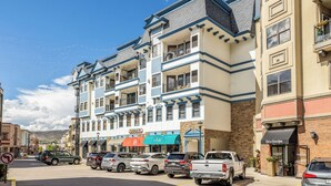 A multi-story hotel with balconies, a variety of shops on the ground floor, and a parking lot with several vehicles. - Riverfront Luxury- Minutes to Skiing+Garage+Walk to shops & restaurants! (Edwards)