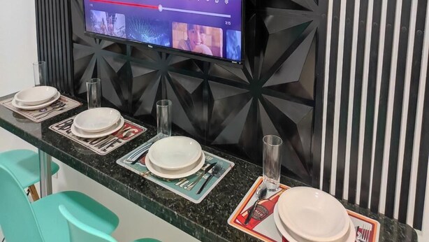 A modern bar area with a flat-screen TV displaying a music video, neatly arranged bar settings, and a geometric wall design. - Ideal Apartamento em Curitiba (Curitiba)