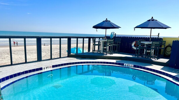 A swimming pool with a view of the beach, a deck area with chairs and umbrellas, and a lifeguard stand.
