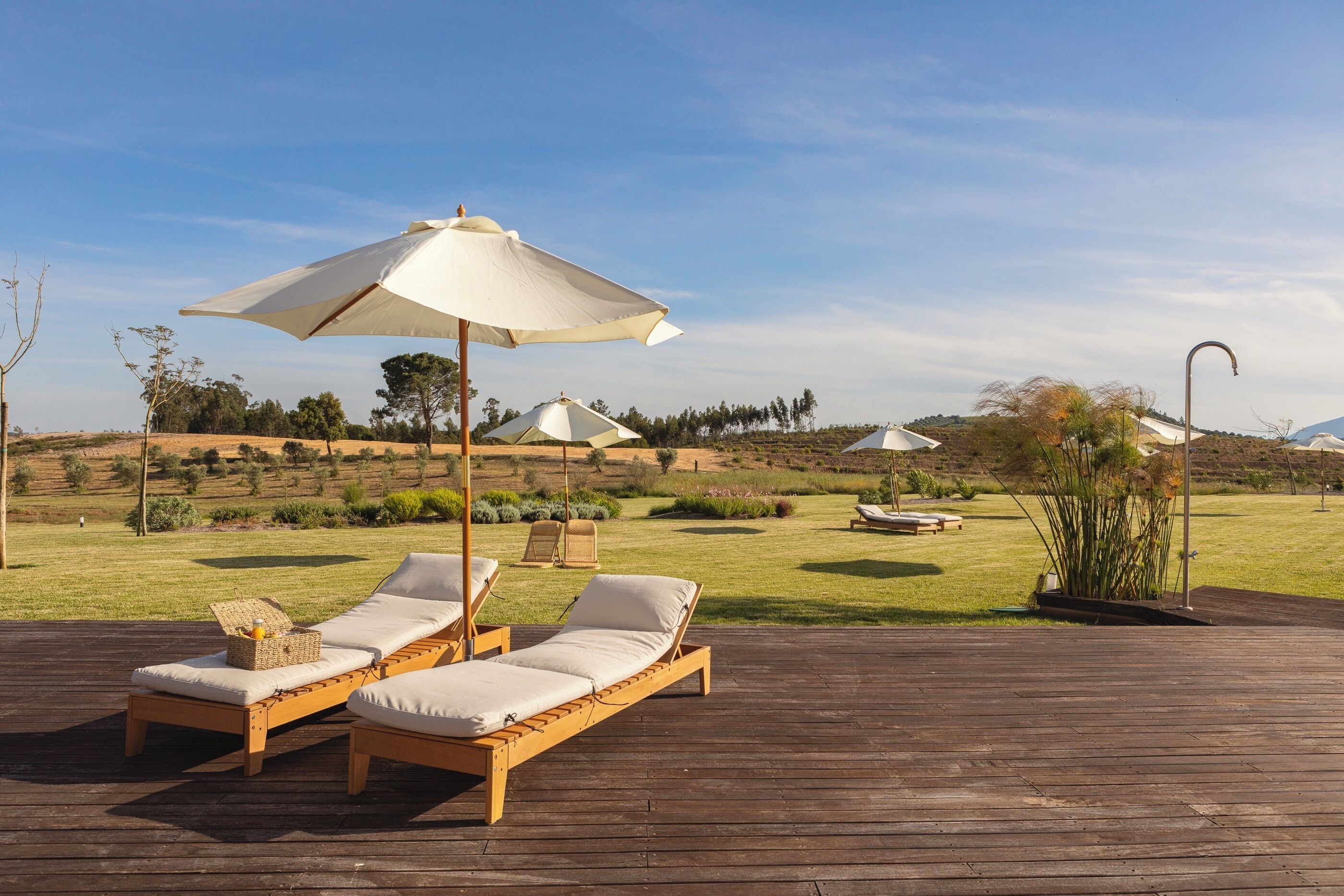 A wooden deck with two sun loungers and a large umbrella, surrounded by a grassy landscape and distant hills.