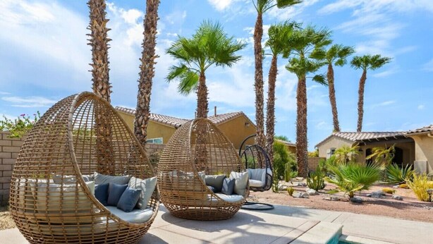 Two wicker swing chairs with cushions by a poolside, surrounded by tall palm trees and a clear sky. - Desert Oasis in Palm Springs/Indio, California (Indio)