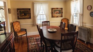 A dining room with a wooden table, six chairs, two windows with white curtains, and two framed paintings on the walls.