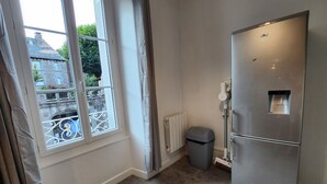 A kitchen with a stainless steel refrigerator, a window with curtains, a radiator, and a trash bin. - Cosy new apartment  (Vic-sur-Cère)