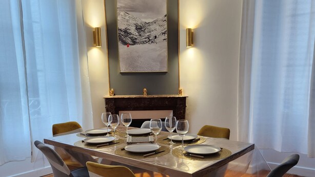 A dining area with a table set for six, surrounded by chairs, a fireplace, and a framed landscape painting. - Cosy new apartment  (Vic-sur-Cère)