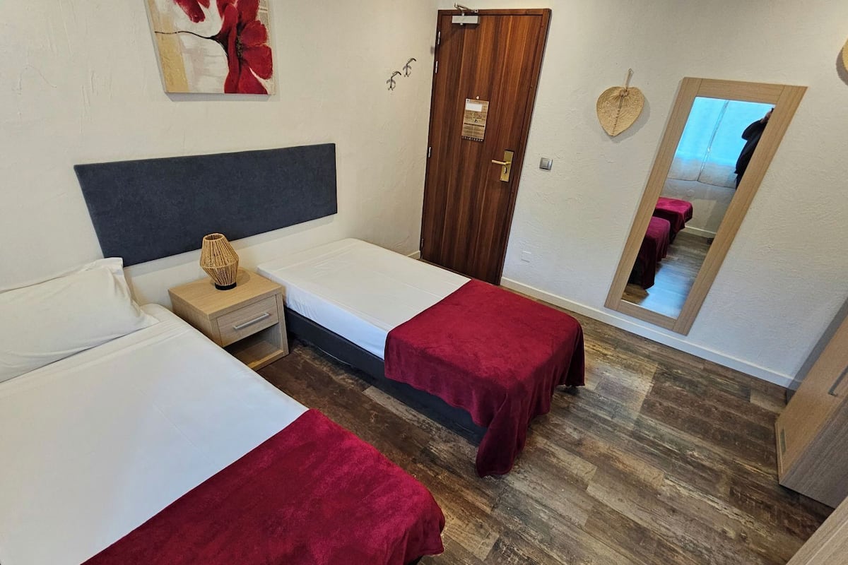 Una habitación con dos camas, una puerta de madera, un espejo y una mesita con una lámpara.