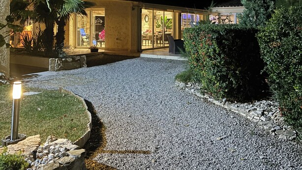 A building with a gravel driveway leading to a glass entrance, surrounded by greenery and a well-lit pathway.