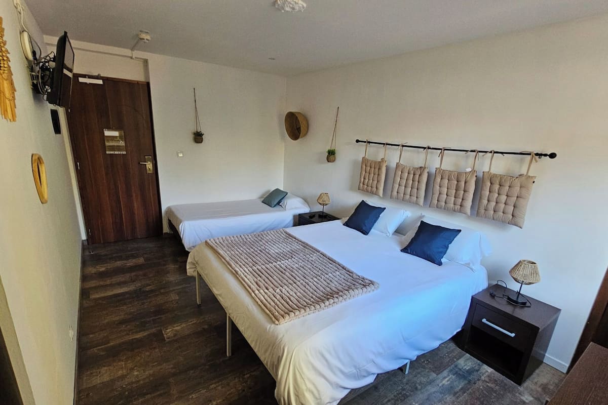 Un dormitorio con dos camas individuales, una cama grande, suelo de madera, una mesita de noche con lámpara y una estantería de pared con cojines.