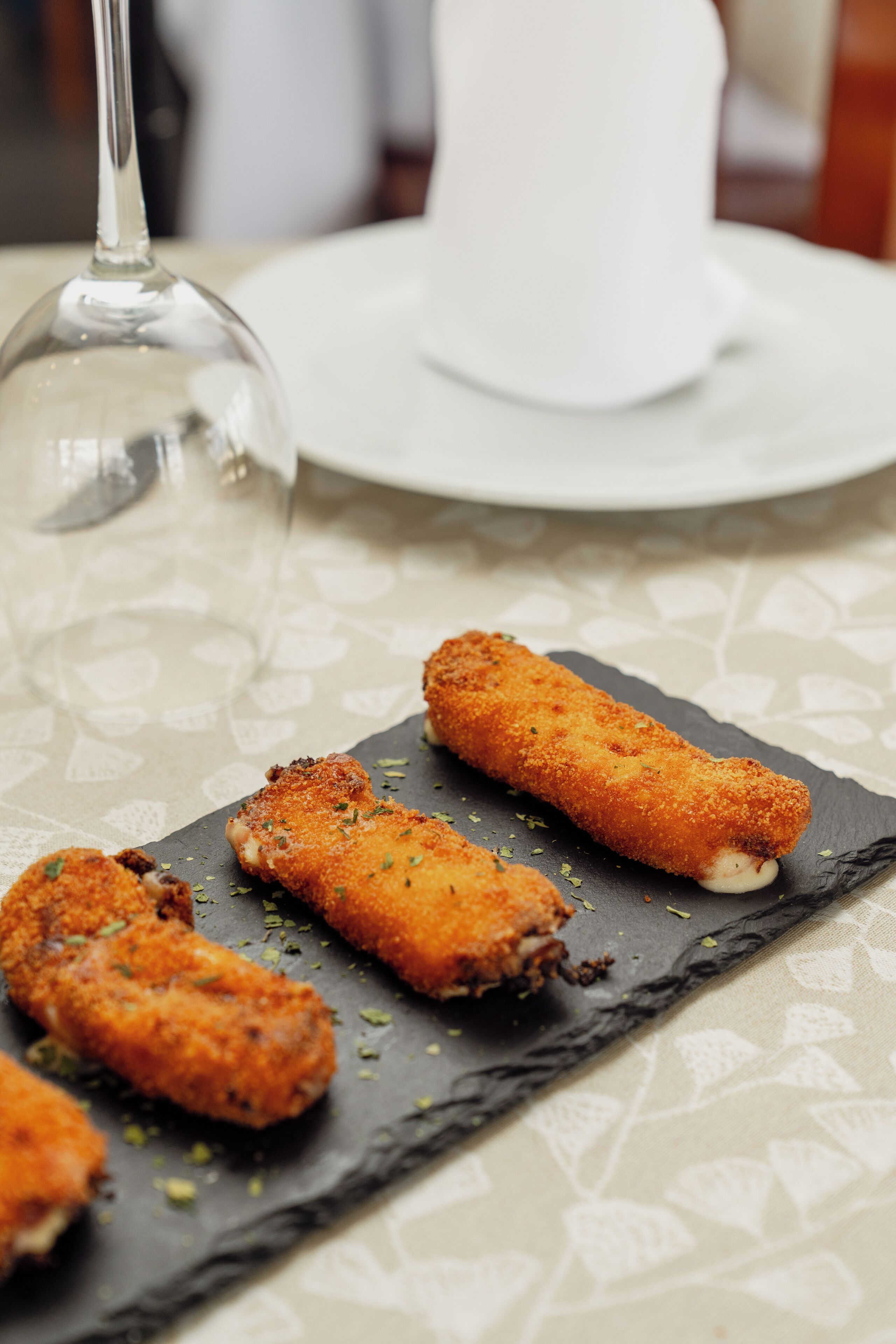 Una tabla de pizarra con tres preparaciones empanizadas y fritas, una copa de vino y un plato blanco con una servilleta sobre una mesa con mantel estampado.