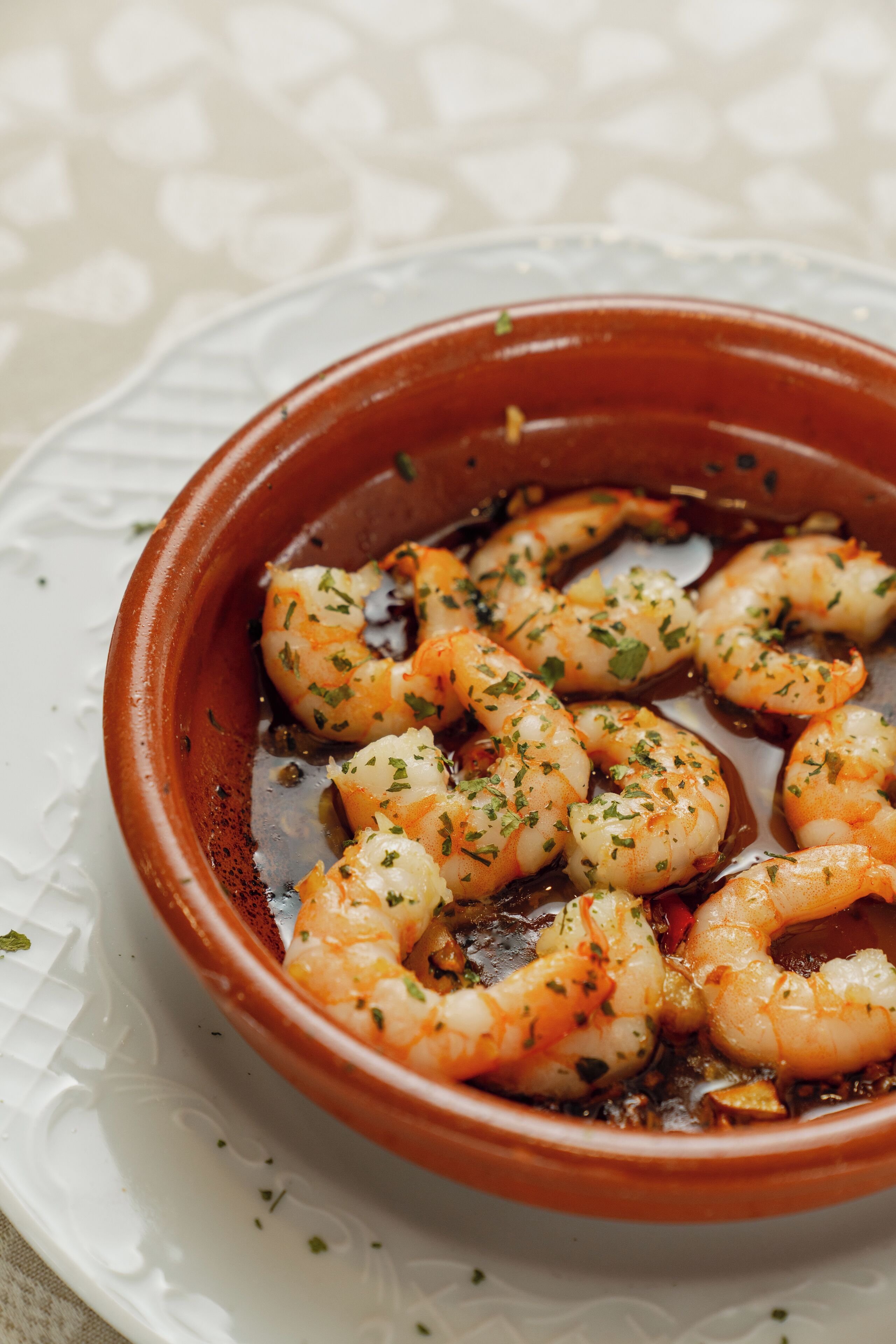 A terracotta dish with cooked shrimp garnished with herbs, served on a white plate with a textured pattern.