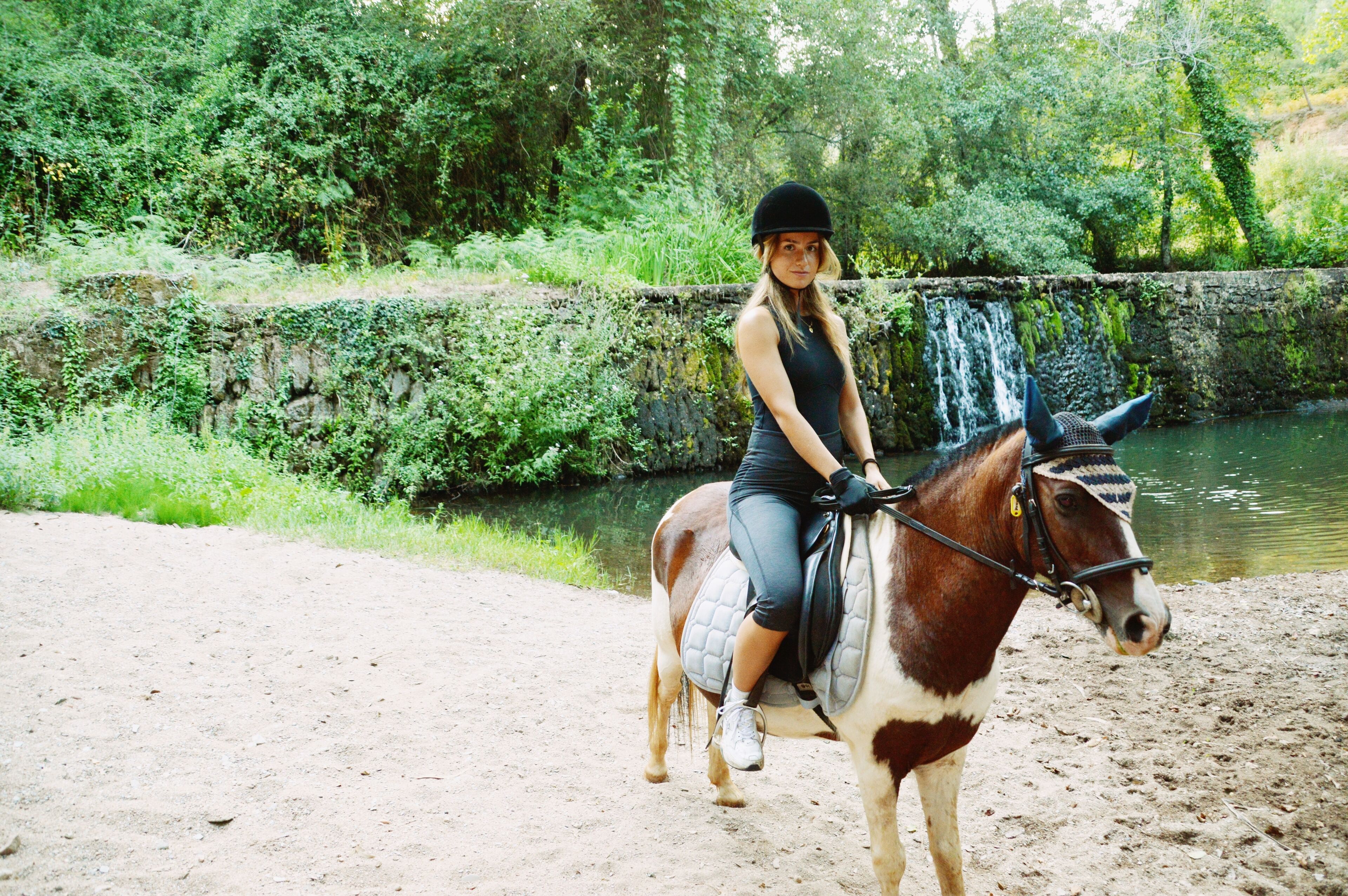 Una persona monta a caballo por un sendero de tierra al lado de un cuerpo de agua, con vegetación al fondo.
