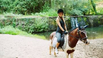 Una persona monta a caballo por un sendero de tierra al lado de un cuerpo de agua, con vegetación al fondo.