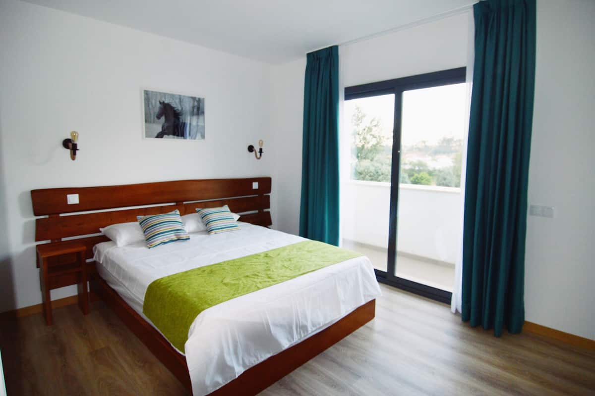 Une chambre avec un lit en bois, du linge de lit blanc, un plaid vert, un tableau au mur et une vue à travers une porte coulissante.