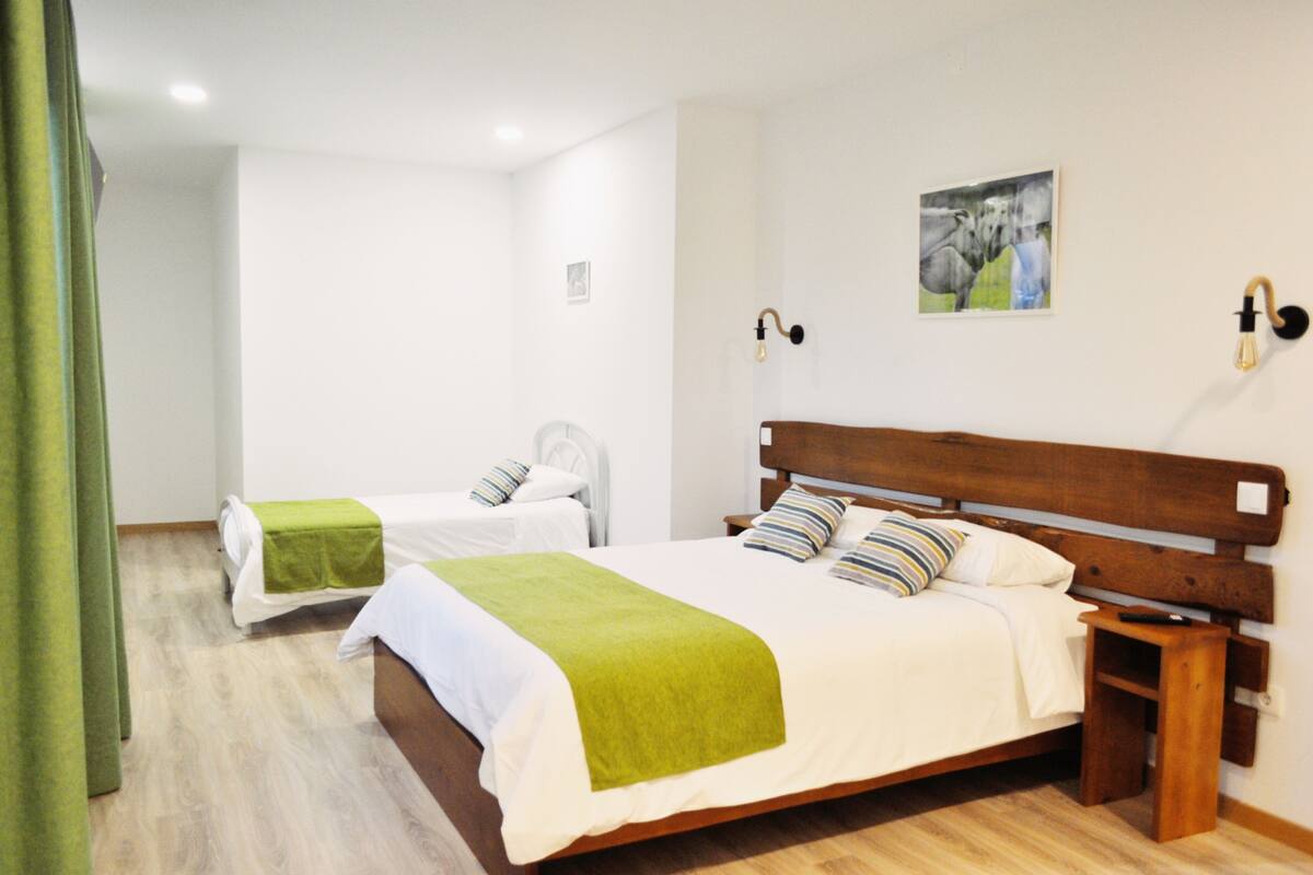 Une chambre d’hôtel avec un grand lit, un lit plus petit, une table de chevet en bois, un luminaire fixé au mur et un tableau accroché au mur.