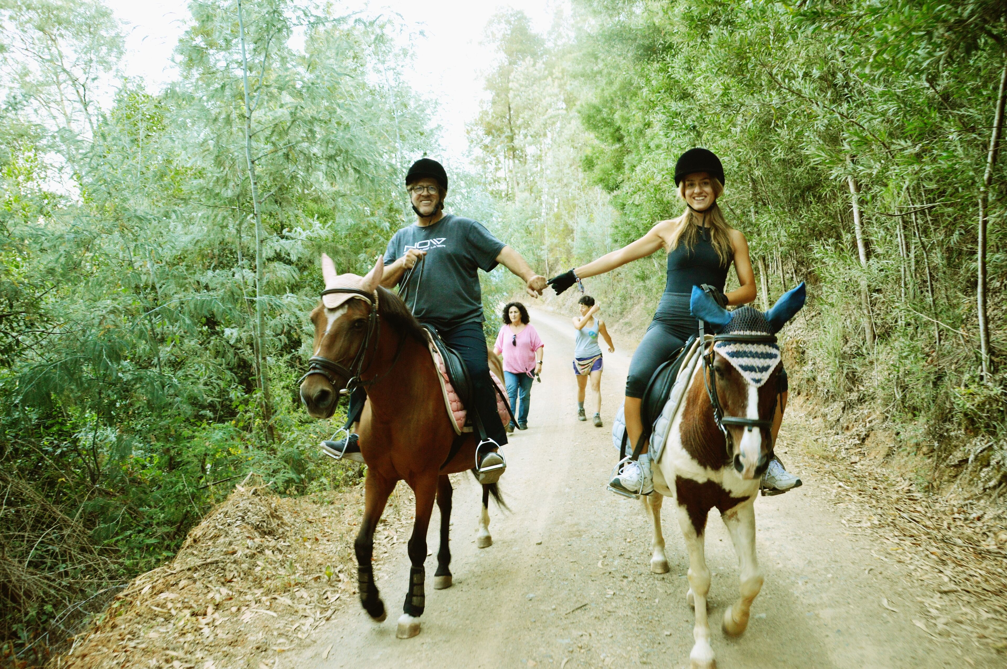 Two people riding horses on a dirt path surrounded by trees.