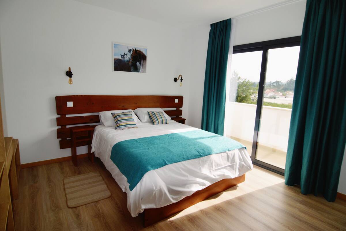 Une chambre avec un lit en bois, une vue dégagée sur l’extérieur grâce à une grande fenêtre et un tableau accroché au mur.