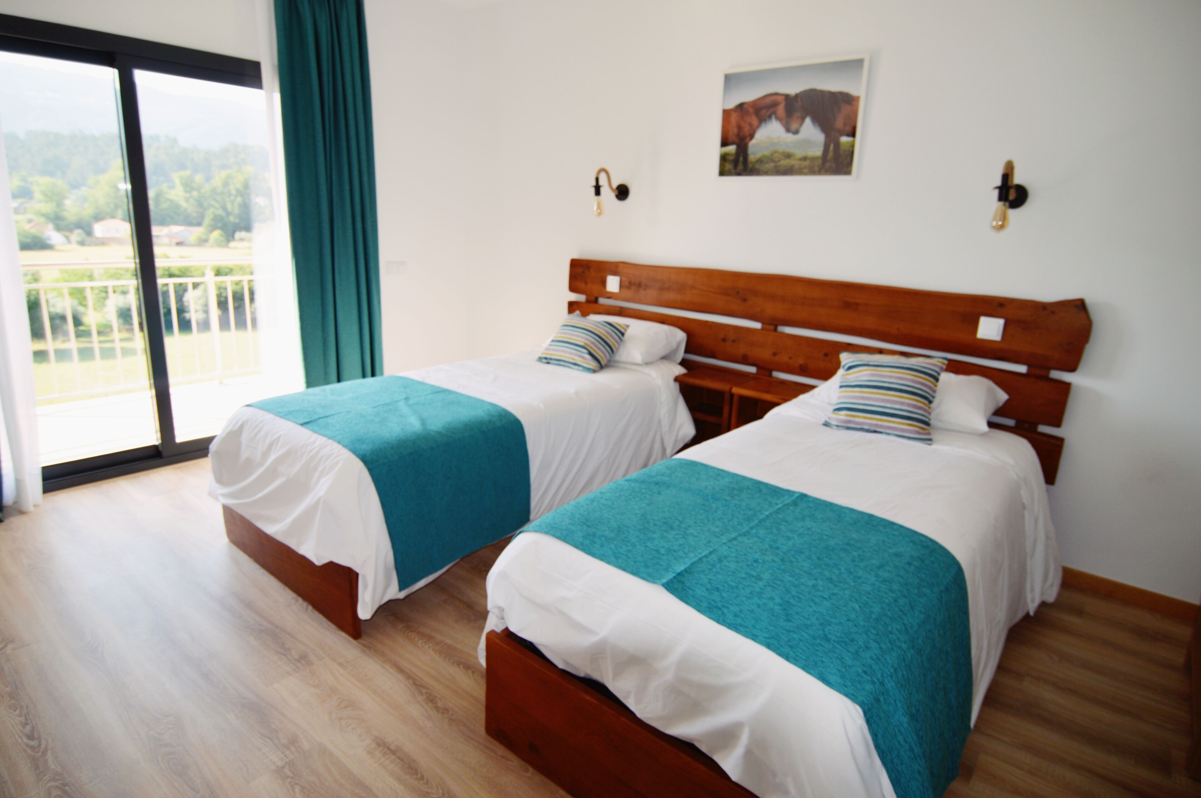Quarto com duas camas, cada uma com cabeceira de madeira e roupa de cama branca, uma janela grande com vista para área verde, e uma fotografia presa na parede.