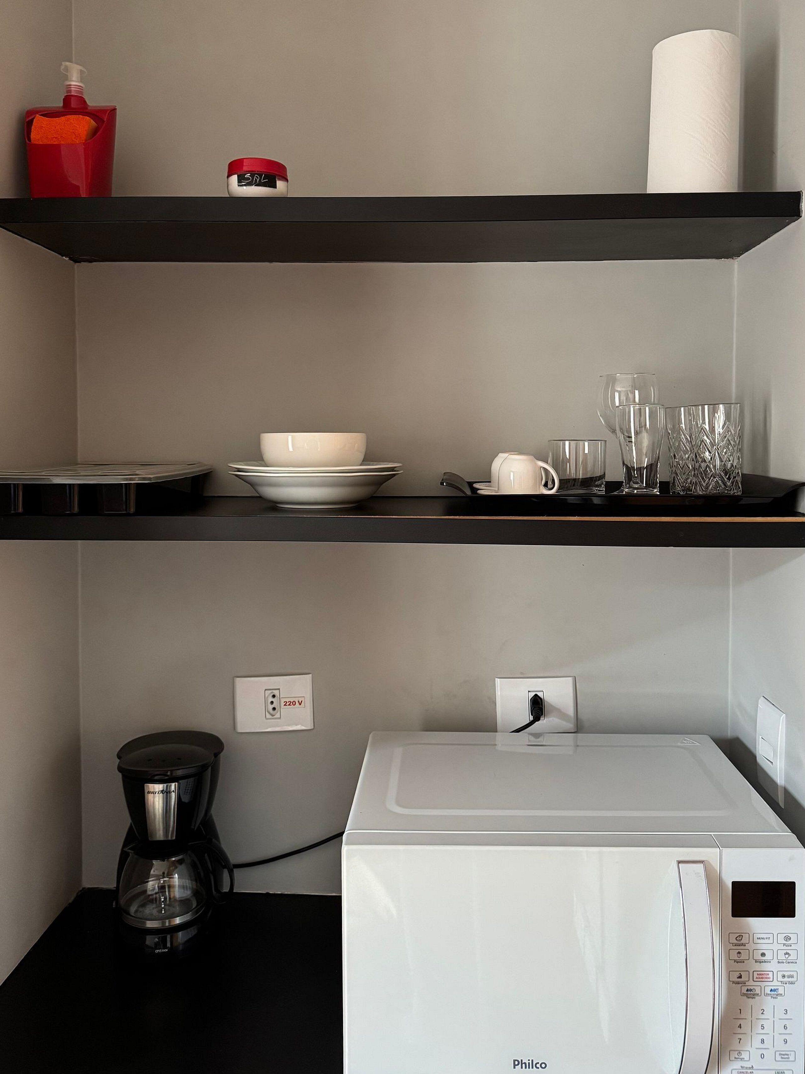 A kitchen with a microwave, coffee maker, and a shelf with dishes and glasses.
